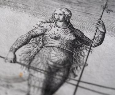 Detail of a printed book shows an illustration of a woman with long hair, elaborate drawings on her body, and holding a spear.